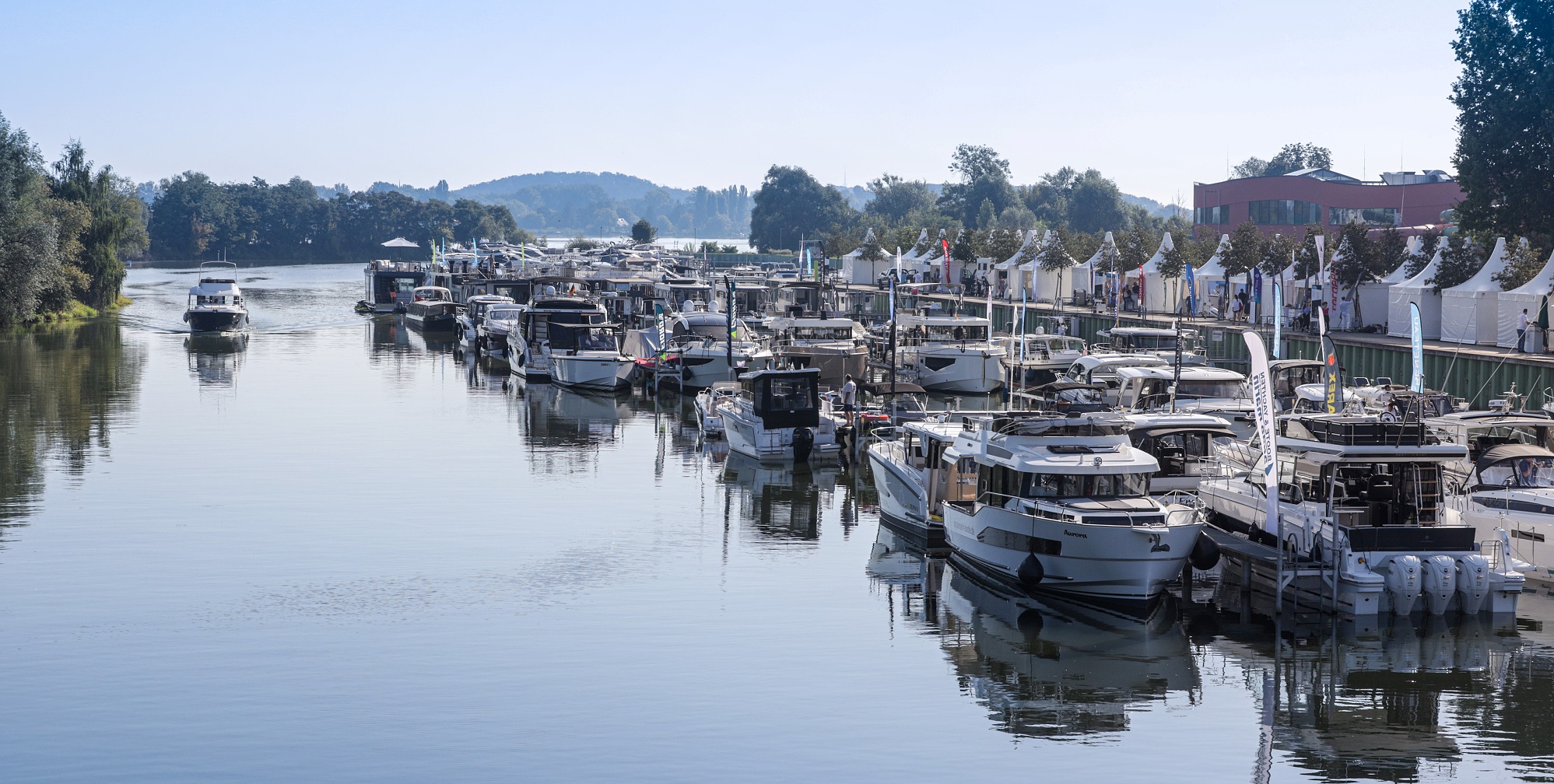 In der Marina Havelauen liegen Boote aller Segmente und Größenklassen an den Stegen. Ein Motorboot fährt in den Marinahafen ein. Der blaue Himmel spiegelt sich auf der Wasseroberfläche. 