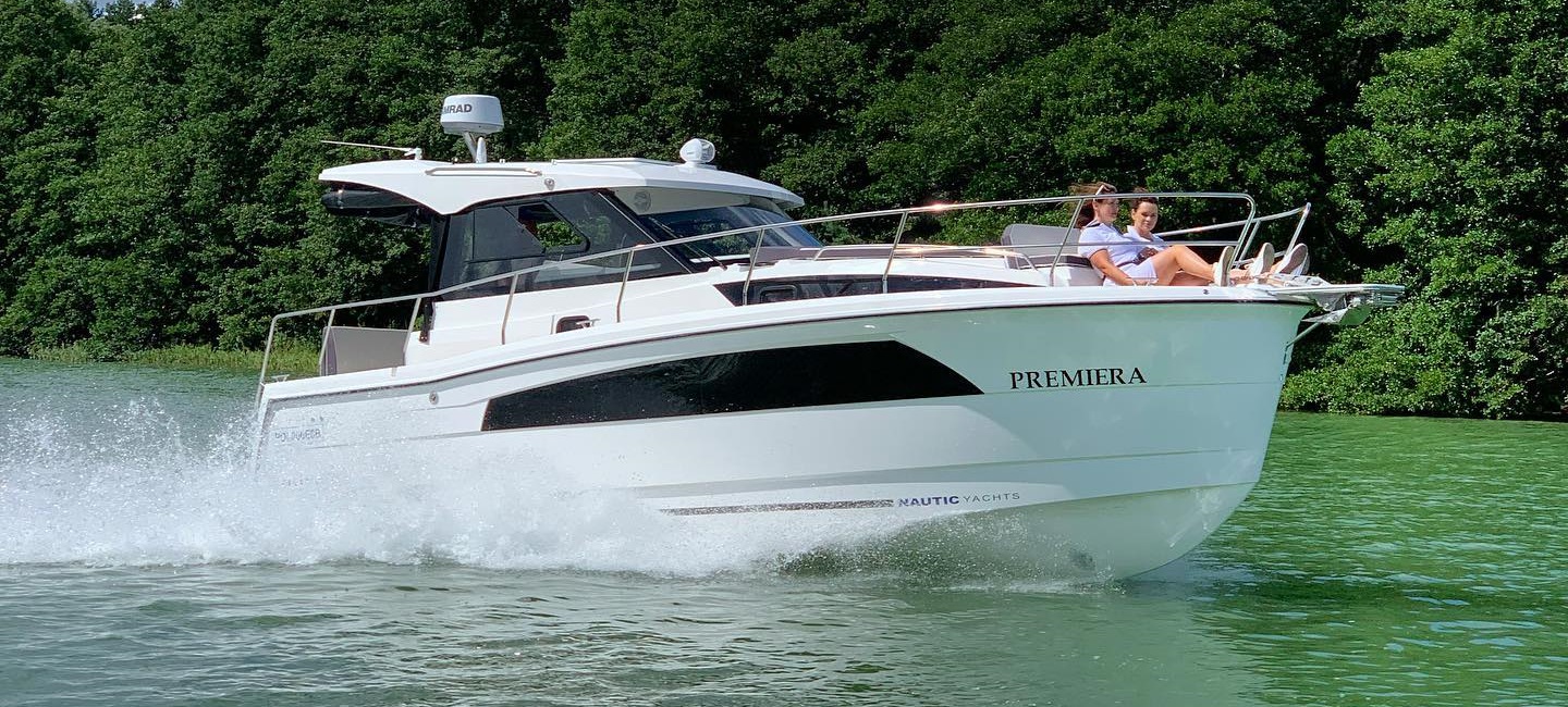 Die Nautic 880 auf dem Wasser. Auf der Bugspitze sitzen entspannt zwei Frauen. Im Hintergrund sieht man am Ufer dicht aneinandergereihte Bäume