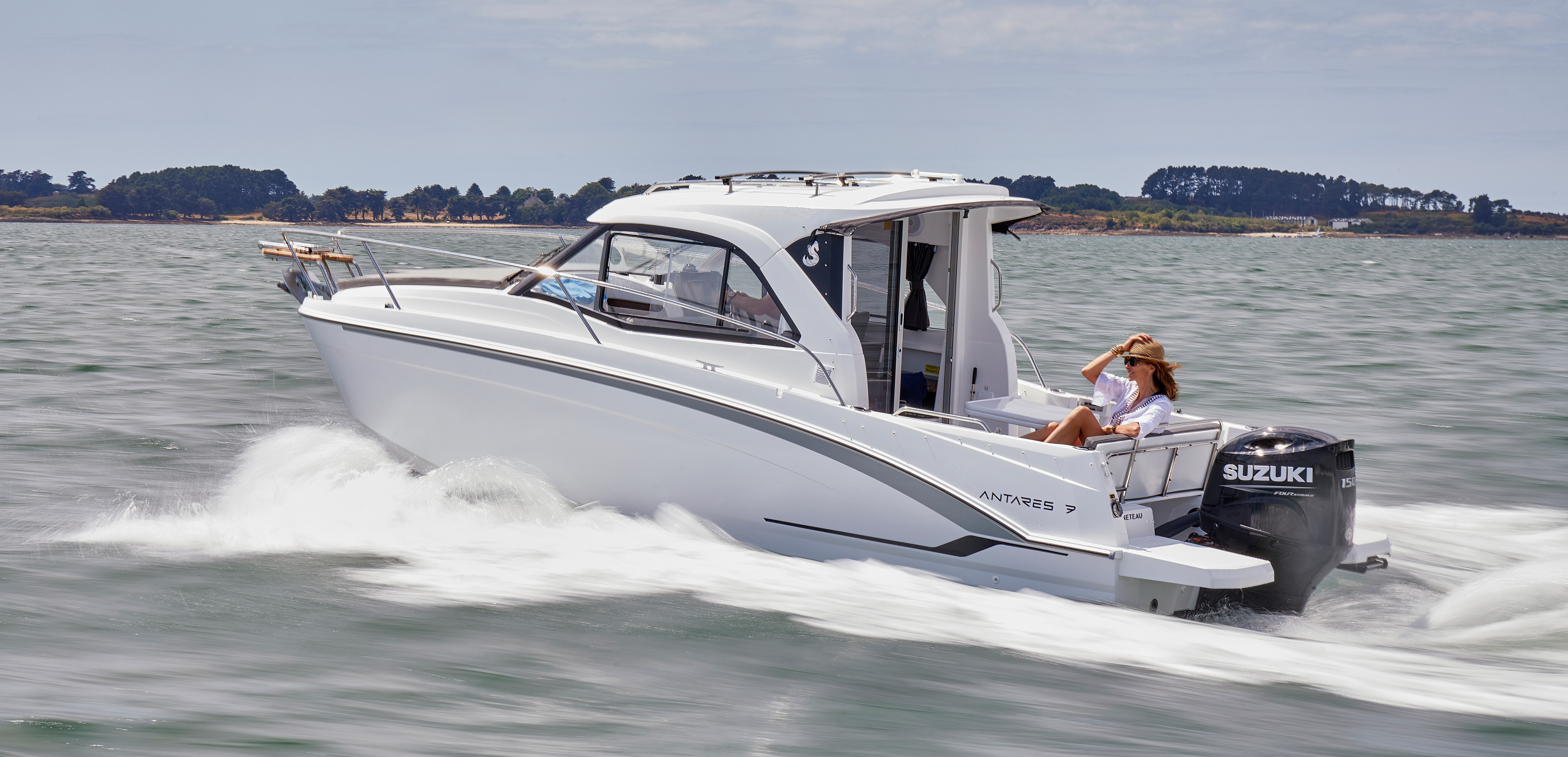 Die weiße Beneteau Antares 7 auf dem Wasser. Im Heck sitzt eine Frau mit Sonnenhut und Sonnenbrille. Im Hintergrund das Ufer mit Bäumen und Büschen.