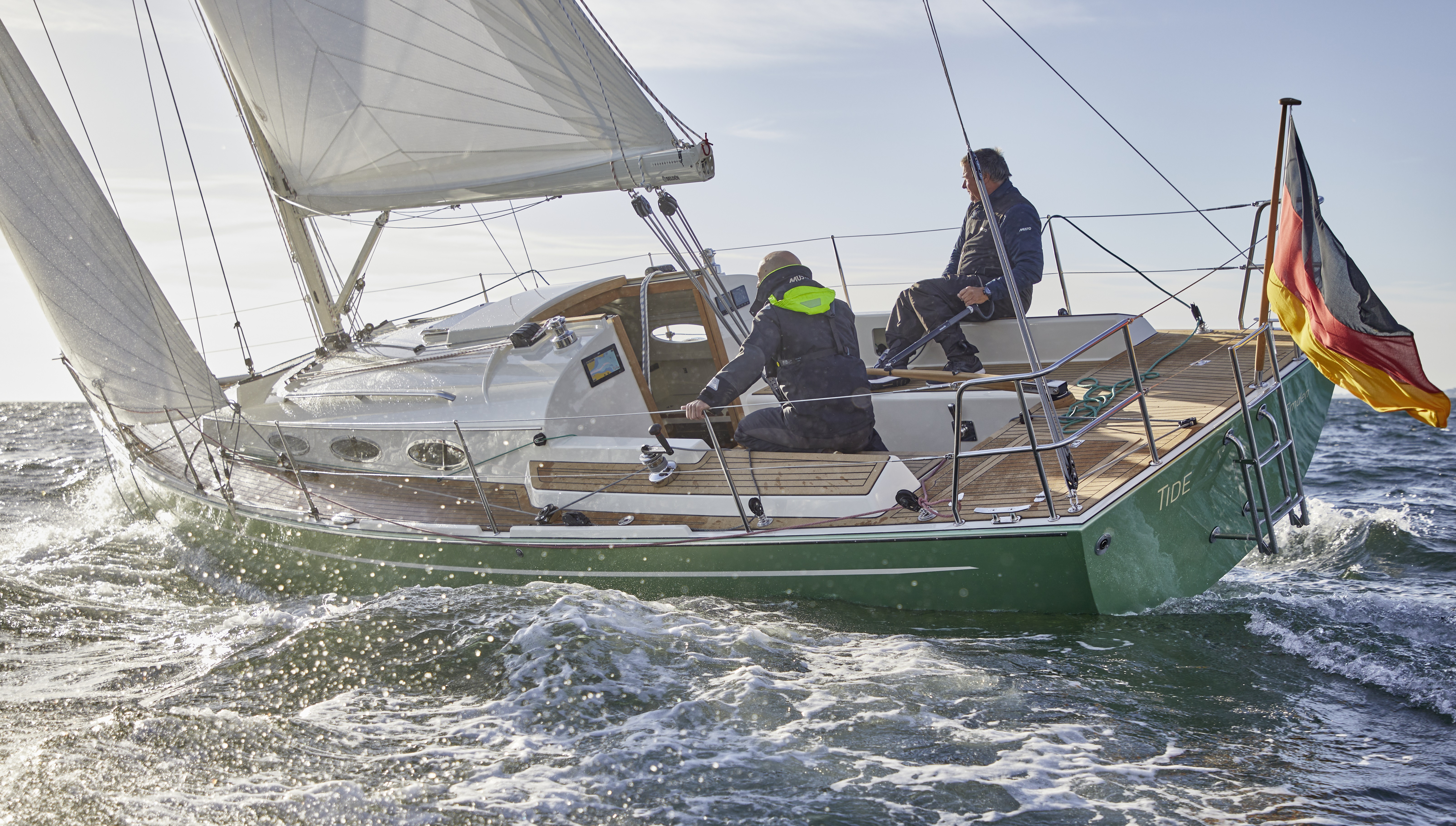 Das Segelboot Tide 36 auf dem Wasser. Zwei Männer in Overalls steuern das schräg im Wasser liegende Boot. Am Heck weht eine Deutschlandfahne.