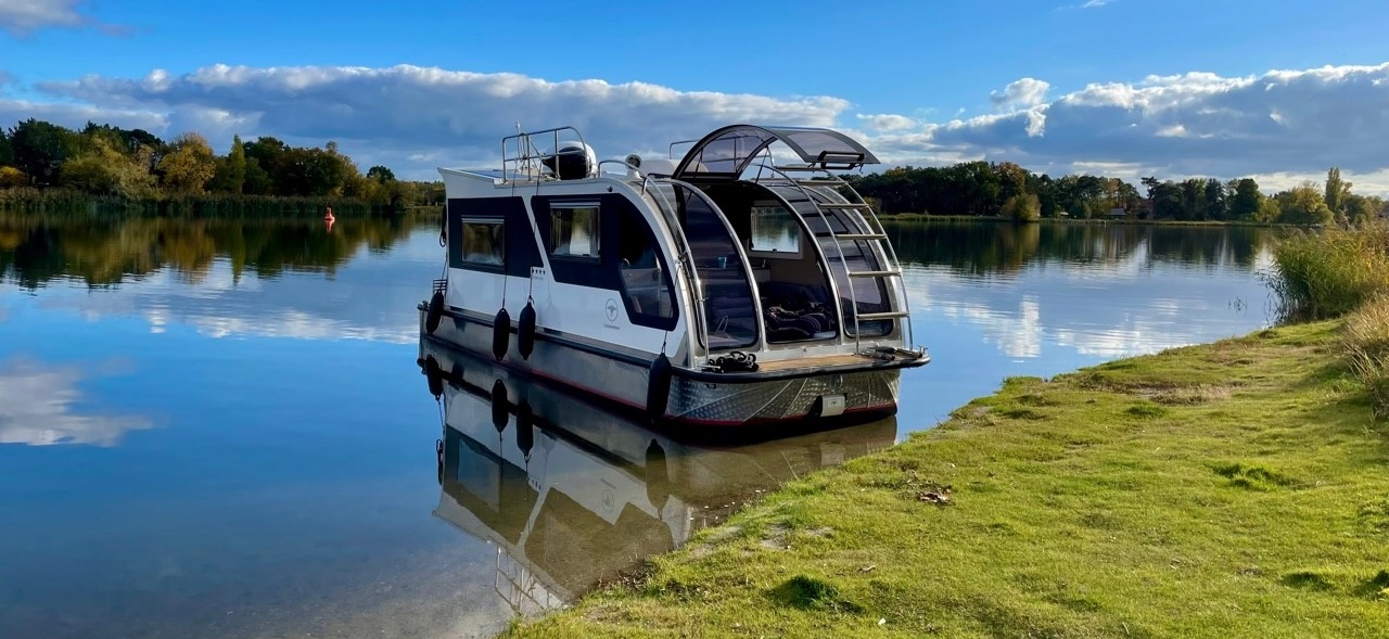 Das Caravanboot Ultimate am Ufer eines Binnengewässers. Die gläserne gewölbte Tür ist nach oben geöffnet. Im Hintergrund das Ufer mit Bäumen und Wolken, die sich im Wasser spiegeln.