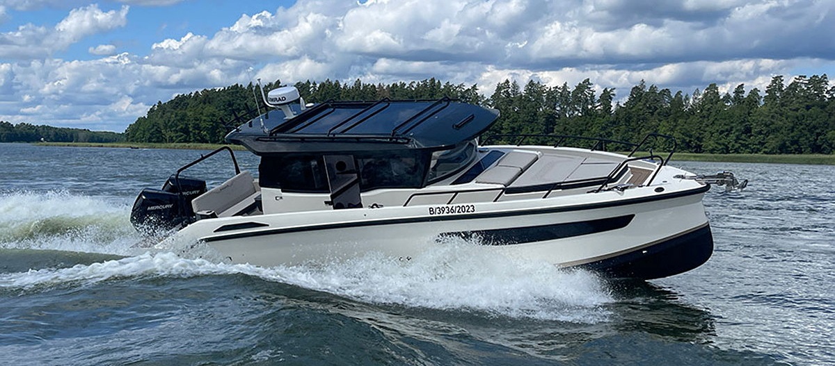 The Navan C30 motorboat – a nine-meter long cabin cruiser with an angular design – sails across a lake. The lake shore can be seen in the background. To the right and left of the boat, the water is sp