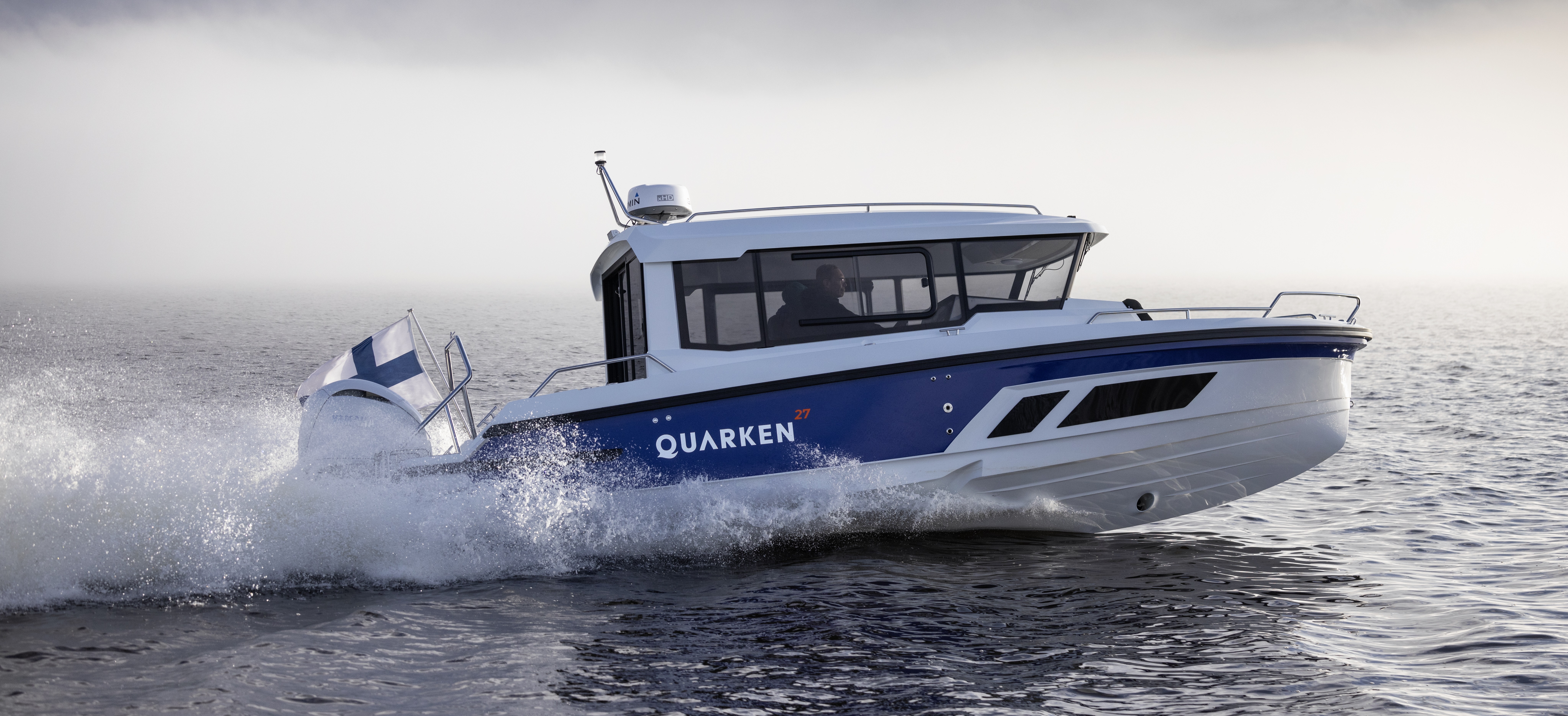 Quarken 27 Cabin motorboat cruises the sea at high speed. The all-weather model has a closed wheelhouse. To the right and left of the boat, the water is splashing. 